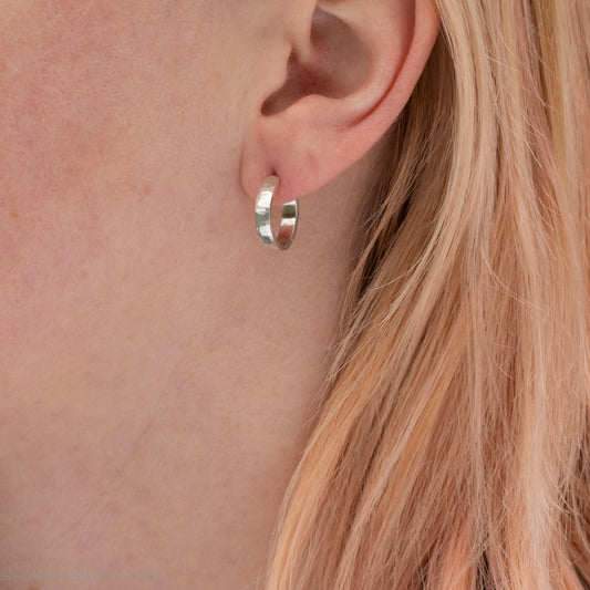A close-up view of a person wearing handcrafted solid silver hoop earrings, 14mm diameter x 3mm wide x 1.5mm thick with a hammered matte finish.