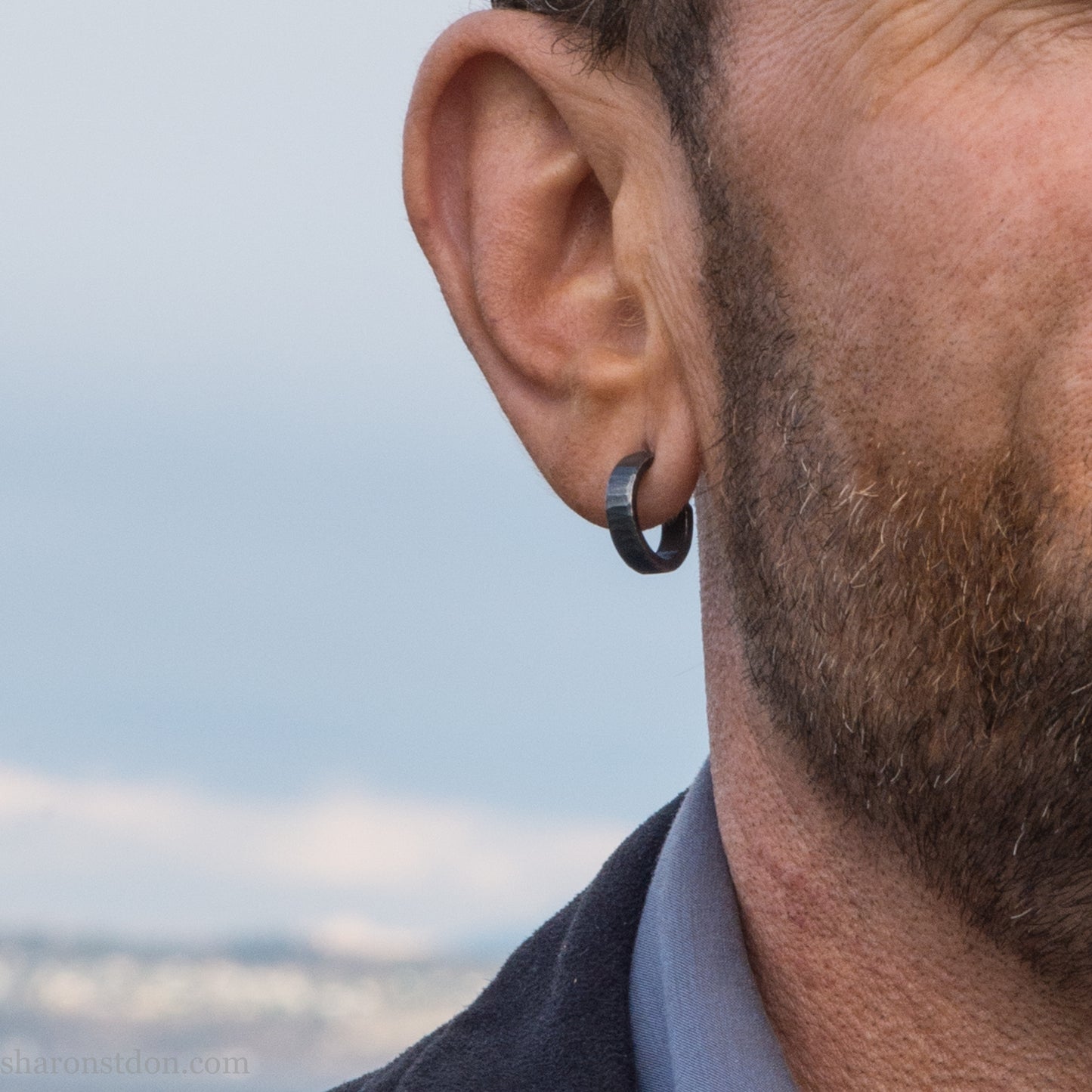 16 x 4mm x 1.5mm thick, 925 sterling silver hoop earrings. Handcrafted in the USA by Sharon Saint Don. Hammered solid silver with an oxidized black finish, silver posts and backs. Worn by a man with fair skin, dark hair and beard.