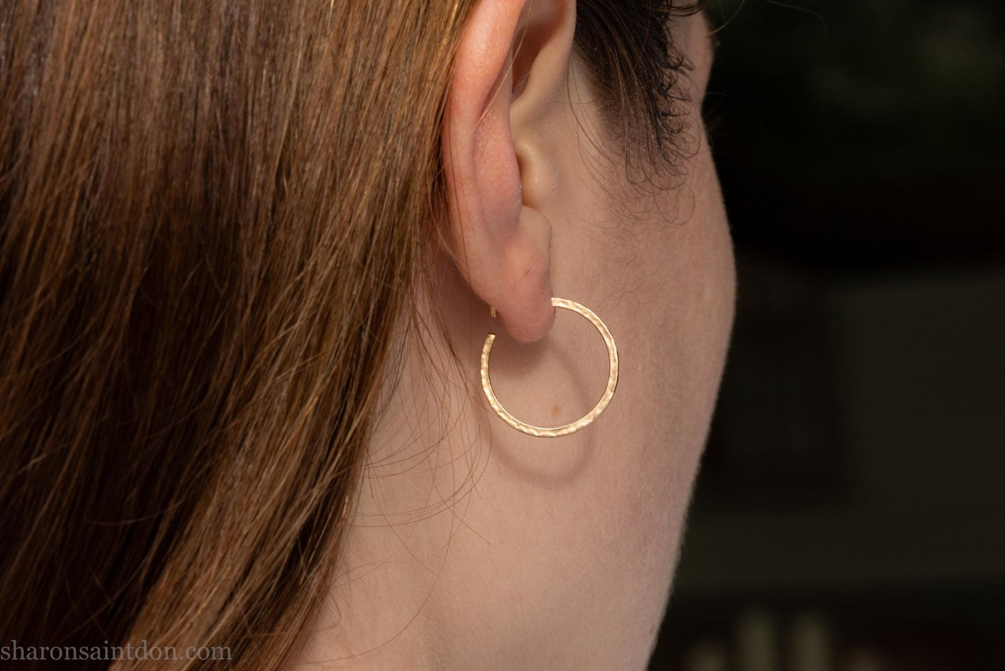 20mm diameter by 1.5mm thick solid 14k yellow gold hoop earrings. Solid 14k gold posts and backs. Handmade by Sharon SaintDon in North America. Hammered from solid gold with a matte finish. Worn by a woman with light skin and brown hair.