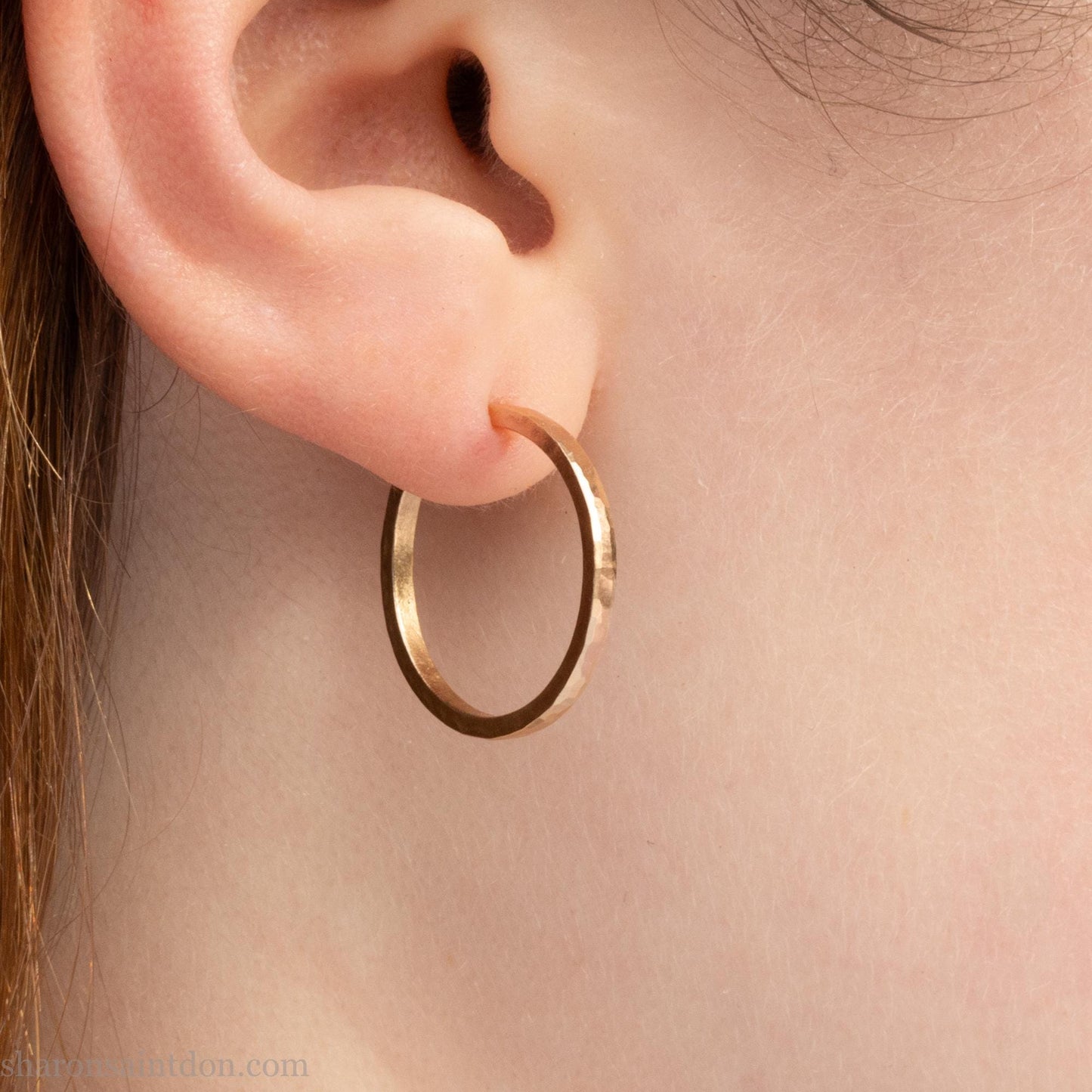 20mm diameter by 1.5mm thick solid 14k yellow gold hoop earrings. Solid 14k gold posts and backs. Handmade by Sharon SaintDon in North America. Hammered from solid gold with a matte finish. Worn by young person with light skin.