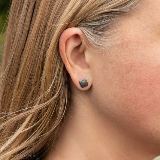 Handcrafted sterling silver square stud earrings with a 2mm canary yellow diamond set in the corner, hammered texture, oxidized black finish, worn by a person.