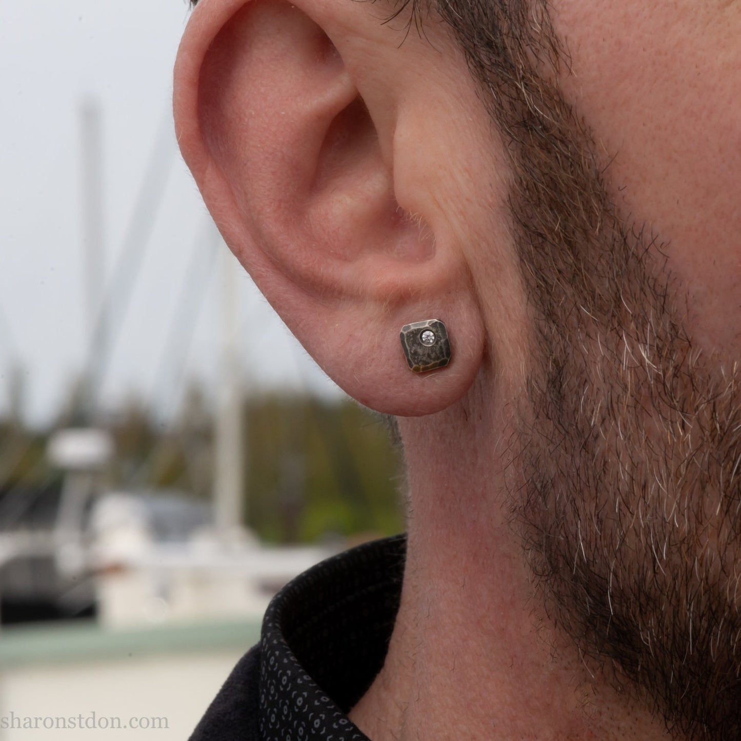 Small 925 sterling silver stud earrings for men with 2mm, imitation diamond gemstone set in the corner. 7mm square, 1.5mm thick with silver posts and backs and hammered, dark, antiqued finish. Handcrafted by Sharon Saint Don in North America. Worn by a man with fair skin, dark hair and beard.