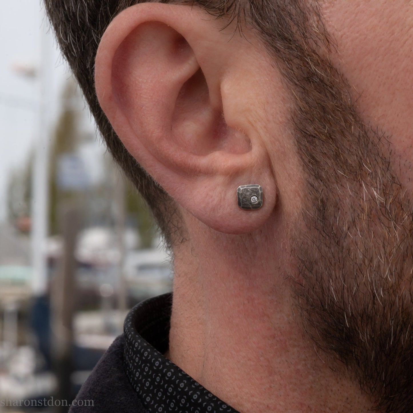 Small 925 sterling silver stud earrings for men with 2mm, imitation diamond gemstone set in the corner. 7mm square, 1.5mm thick with silver posts and backs and hammered, dark, antiqued finish. Handcrafted by Sharon Saint Don in North America. Worn by a man with fair skin, dark hair and beard.