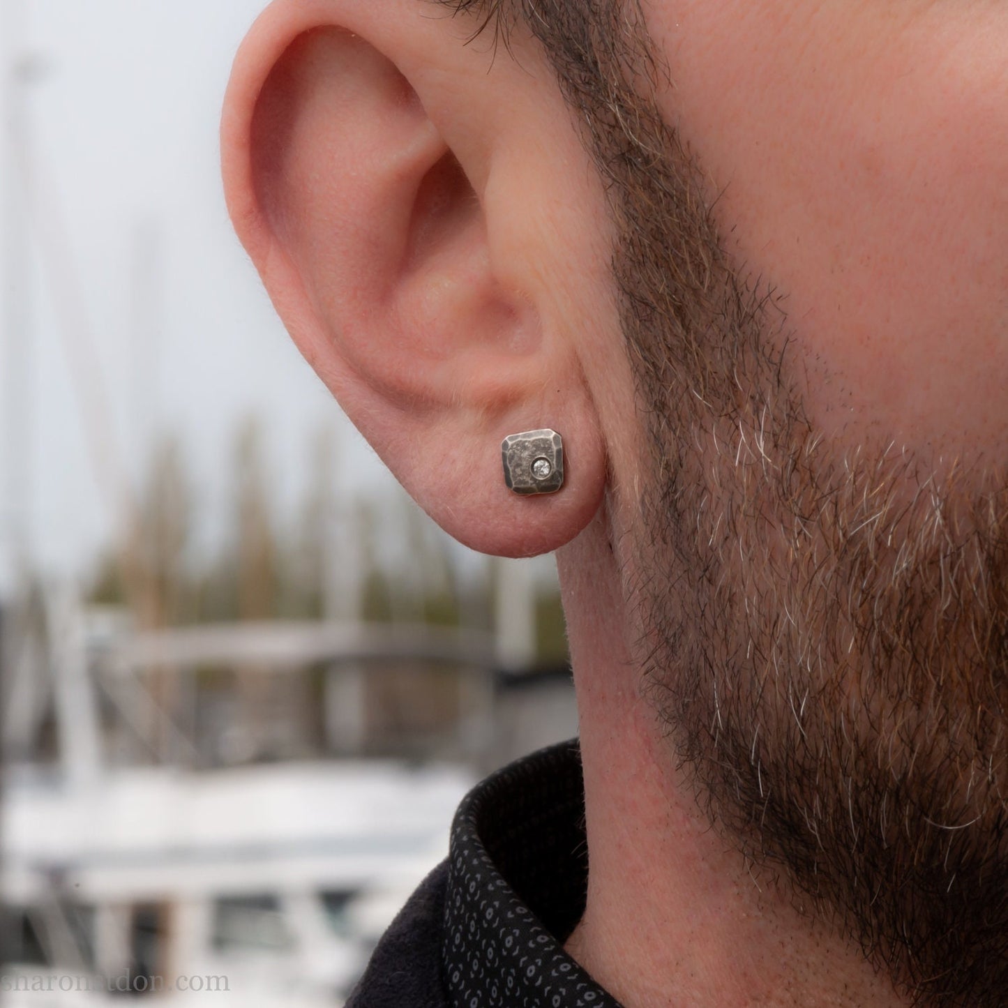 Small 925 sterling silver stud earrings for men with 2mm, imitation diamond gemstone set in the corner. 7mm square, 1.5mm thick with silver posts and backs and hammered, dark, antiqued finish. Handcrafted by Sharon Saint Don in North America. Worn by a man with fair skin, dark hair and beard.