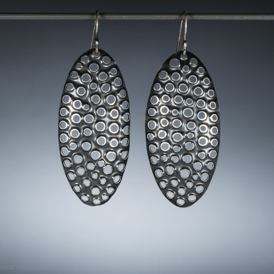 925 sterling silver dangle earrings, 50mm tall ovals with mesh holes punched by hand, and handmade ear wire french hooks. Oxidized black, dark patina rubbed back to shiny white shiny raised circles around the holes like little craters.