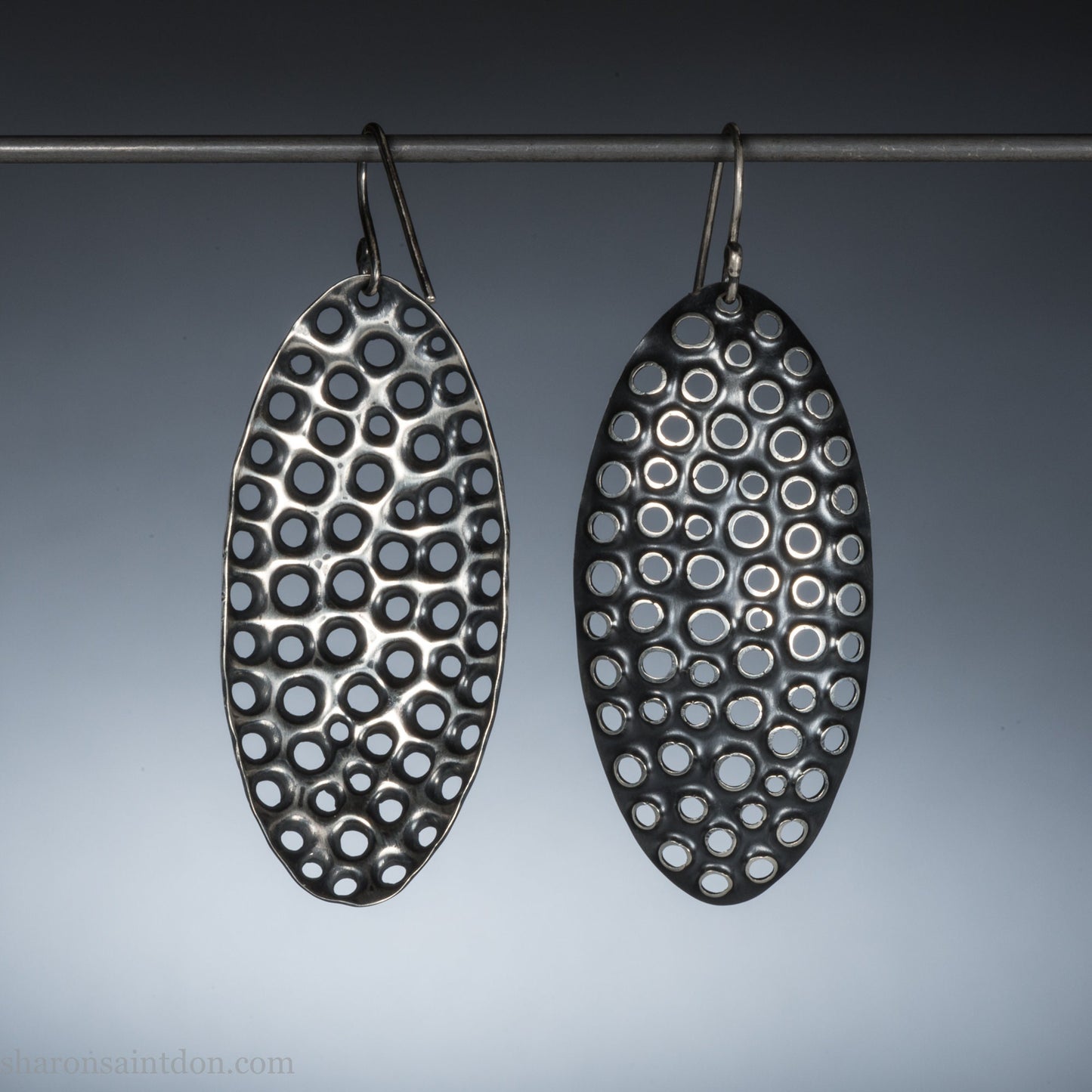 925 sterling silver dangle earrings, 50mm tall ovals with mesh holes punched by hand, and handmade ear wire french hooks. Oxidized black, dark patina rubbed back to shiny white shiny raised circles around the holes like little craters.