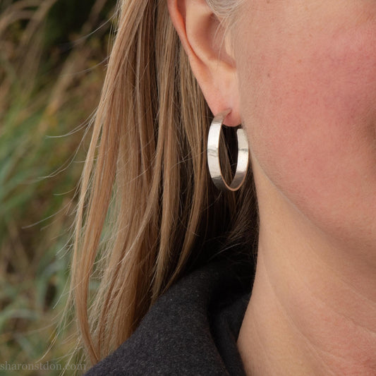 30mm diameter x 4mm wide x 1mm thick solid sterling silver hoop earrings. Handmade in the Pacific Northwest of the USA by Sharon Saint Don. Shiny matte brushed finish, solid silver posts and backs. Pictured on a woman with blonde long hair and fair skin.