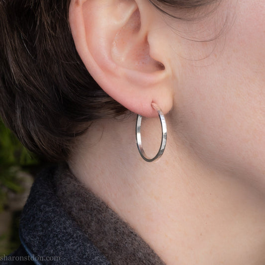 Hammered solid silver hoop earrings, handmade in the USA by Sharon Saint Don. Earrings are worn by a person with fair skin and short brown hair. 25mm diameter, 2mm wide, 1.5mm thick. solid silver posts and backs. Lightweight and comfortable for daily wear with a matte, brushed, shiny finish.