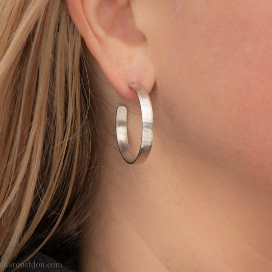 30mm diameter x 4mm wide x 1mm thick solid sterling silver hoop earrings. Handmade in the Pacific Northwest of the USA by Sharon Saint Don. Shiny matte brushed finish, solid silver posts and backs. Pictured on a woman with blonde long hair and fair skin.