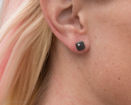 Handcrafted 925 sterling silver stud earrings with moissanite, lab grown, alternative diamond gemstones. 7mm square, 1.5mm thick, solid hammered silver with oxidized black finish. 2mm round moissanite is set in the corner, posts and backs are sterling silver. Worn by a woman with light skin and blonde hair.