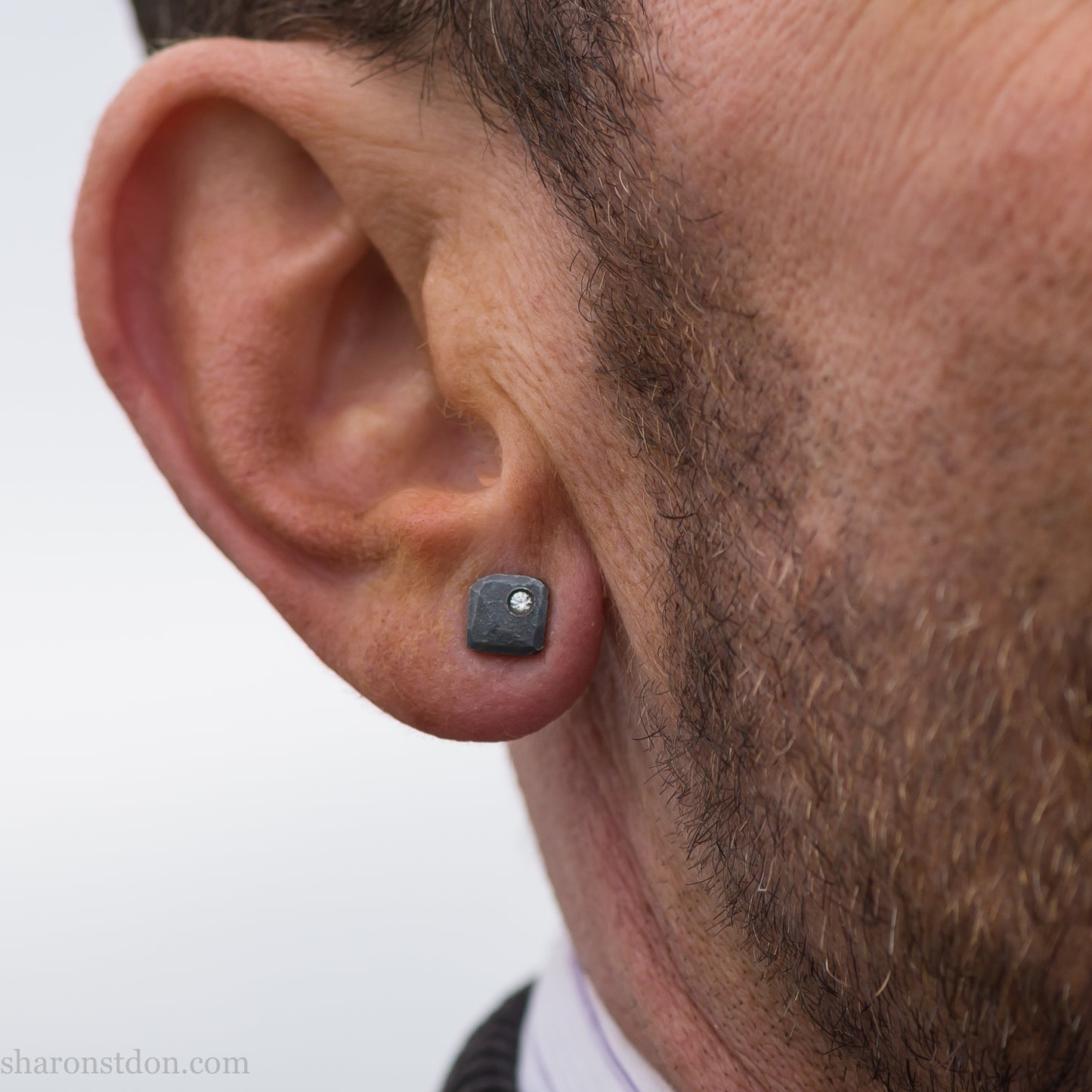 Handcrafted 925 sterling silver stud earrings with moissanite, lab grown, alternative diamond gemstones. 7mm square, 1.5mm thick, solid hammered silver with oxidized black finish. 2mm round moissanite is set in the corner, posts and backs are sterling silver. Worn by a man with light skin, brown hair and beard.