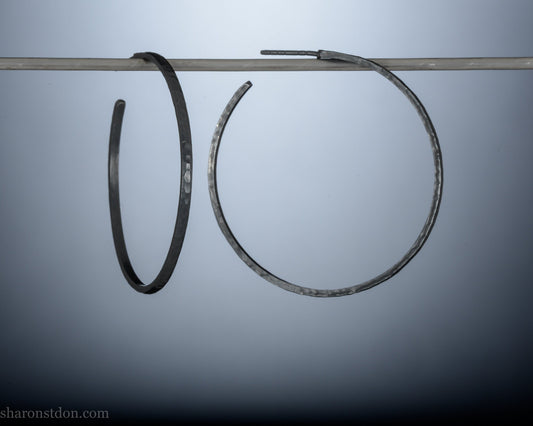 Black 925 sterling silver hoop earrings, 40mm diameter, 2mm wide, 1mm thick. Hammered solid silver with silver posts and backs, worn by a person.
