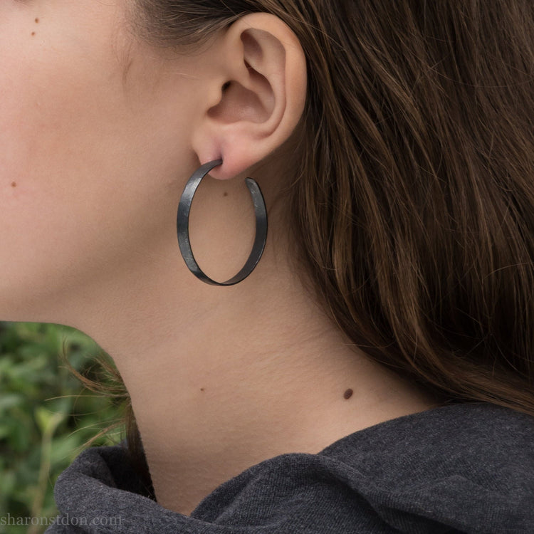 40 mm diameter, 5mm wide, oxidized black sterling silver round hammered hoop earring, close-up on model’s ear