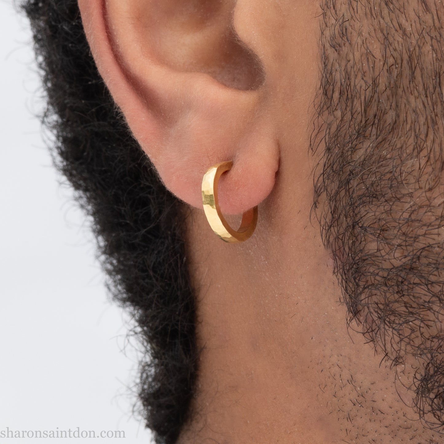 Hammered solid 18k yellow gold hoop earrings. 18mm diameter, 2mm wide, with a matte, brushed finish. Solid 18k gold posts and locking backs. Handmade by Sharon SaintDon in North America. Worn in photo by a young man with dark skin and black hair.