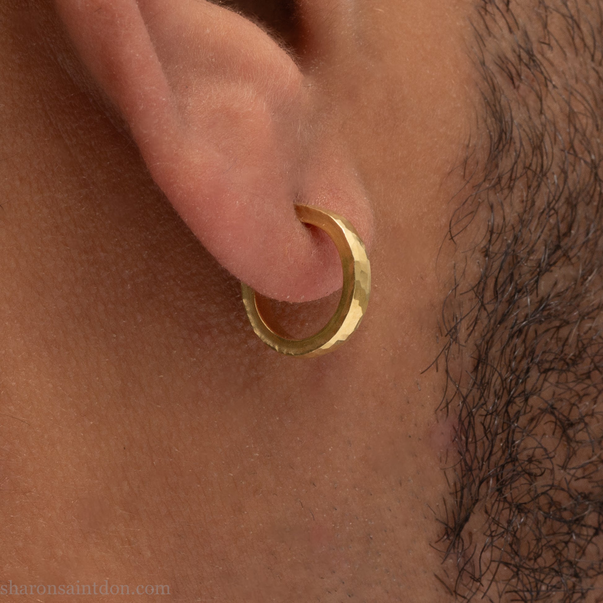 Hammered solid 18k yellow gold hoop earrings. 18mm diameter, 2mm wide, with a matte, brushed finish. Solid 18k gold posts and locking backs. Handmade by Sharon SaintDon in North America. Worn in photo by a young man with dark skin and black hair.