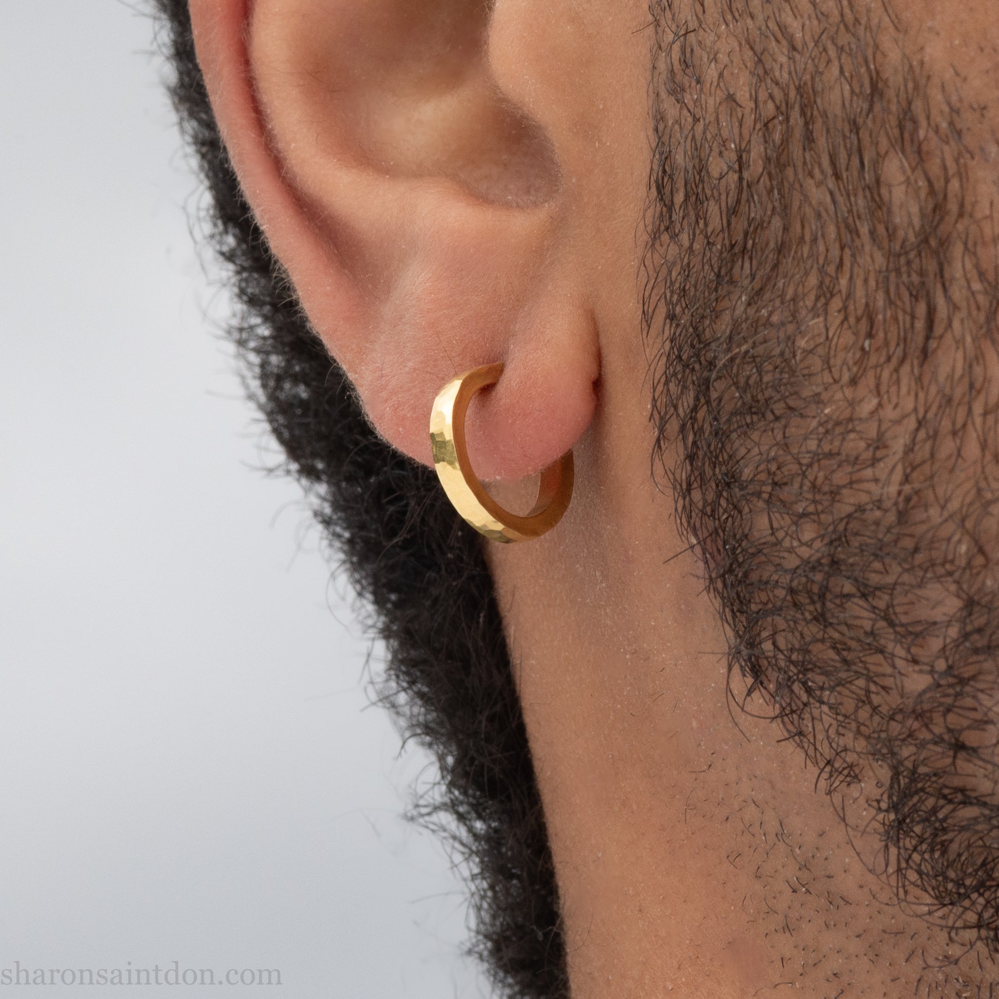 Hammered solid 18k yellow gold hoop earrings. 18mm diameter, 2mm wide, with a matte, brushed finish. Solid 18k gold posts and locking backs. Handmade by Sharon SaintDon in North America. Worn in photo by a young man with dark skin and black hair.