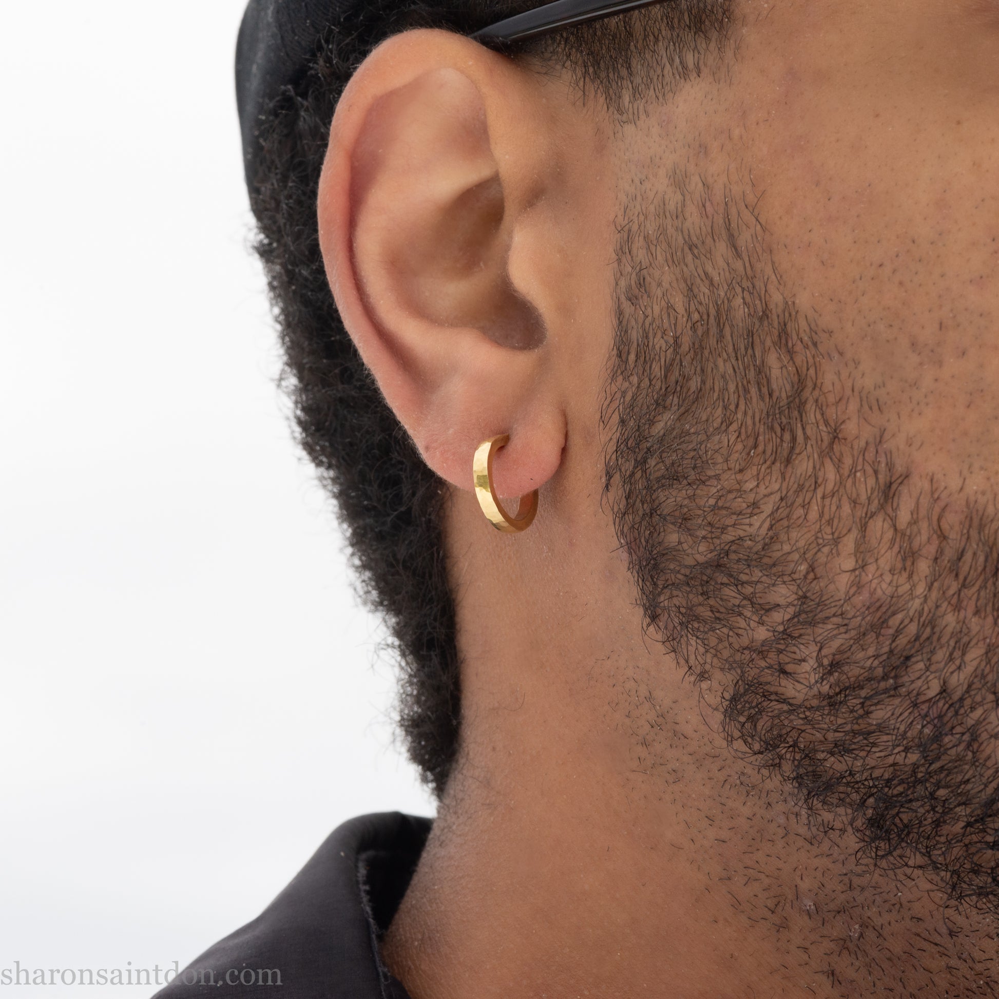 Hammered solid 18k yellow gold hoop earrings. 18mm diameter, 2mm wide, with a matte, brushed finish. Solid 18k gold posts and locking backs. Handmade by Sharon SaintDon in North America. Worn in photo by a young man with dark skin and black hair.