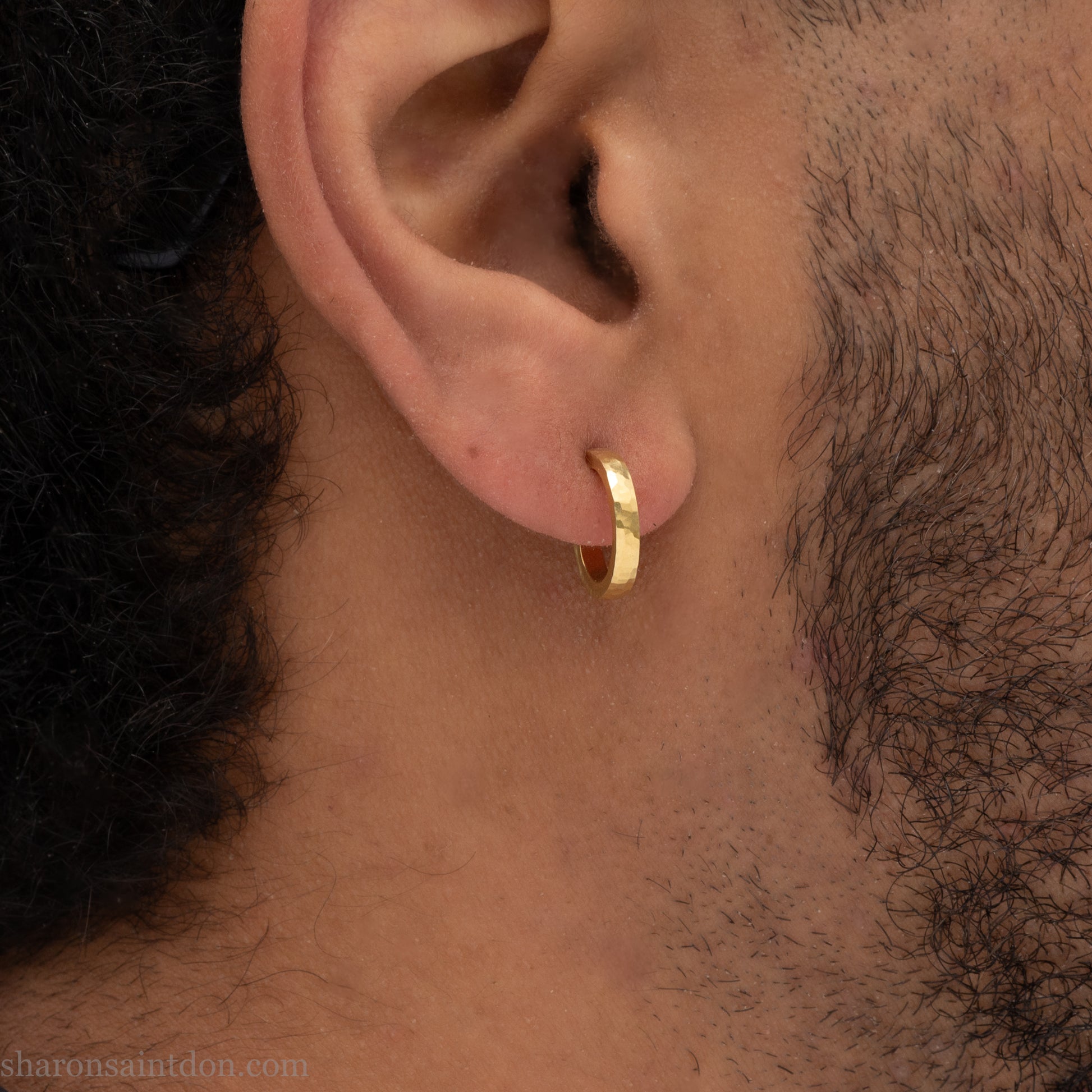 Hammered solid 18k yellow gold hoop earrings. 18mm diameter, 2mm wide, with a matte, brushed finish. Solid 18k gold posts and locking backs. Handmade by Sharon SaintDon in North America. Worn in photo by a young man with dark skin and black hair.
