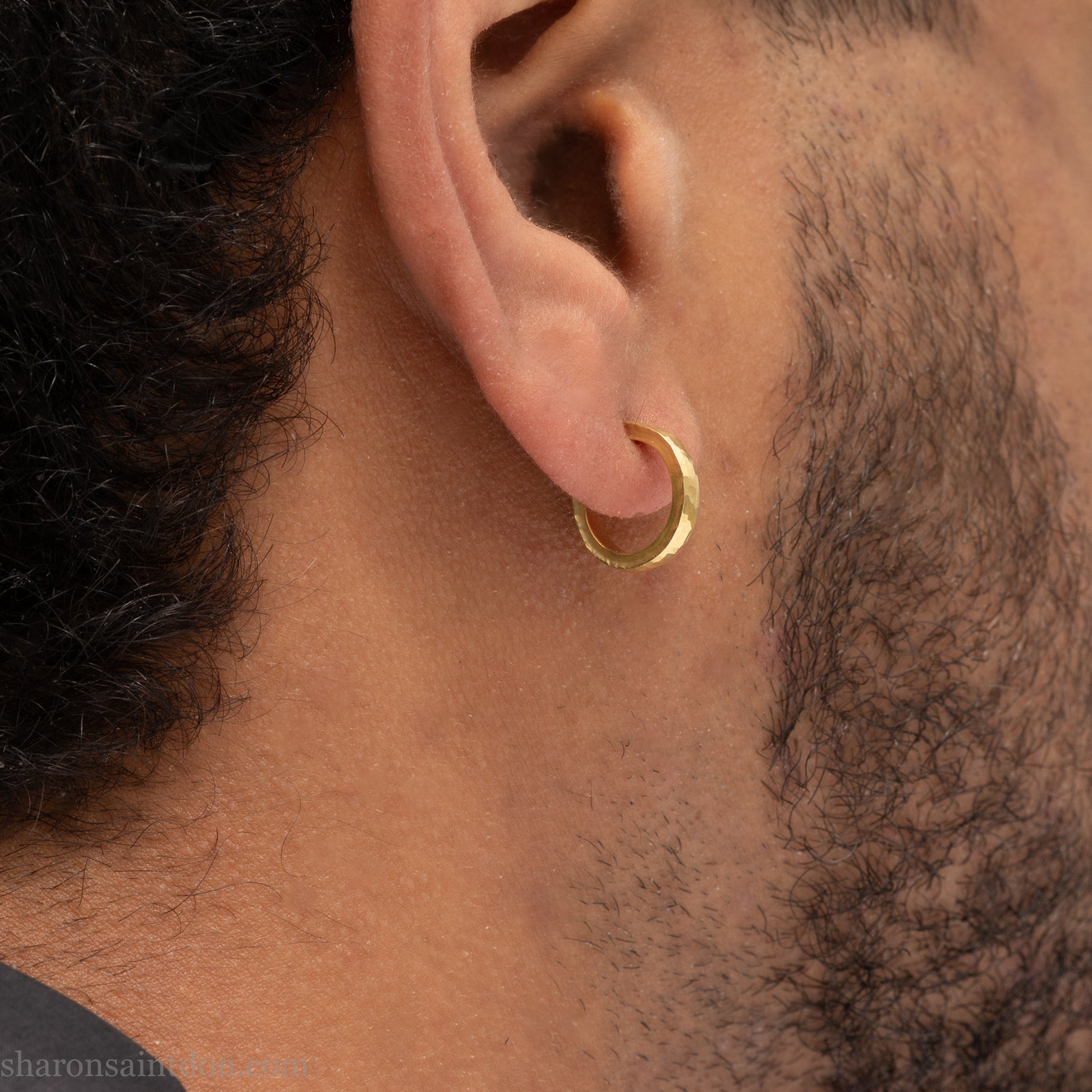Hammered solid 18k yellow gold hoop earrings. 18mm diameter, 2mm wide, with a matte, brushed finish. Solid 18k gold posts and locking backs. Handmade by Sharon SaintDon in North America. Worn in photo by a young man with dark skin and black hair.