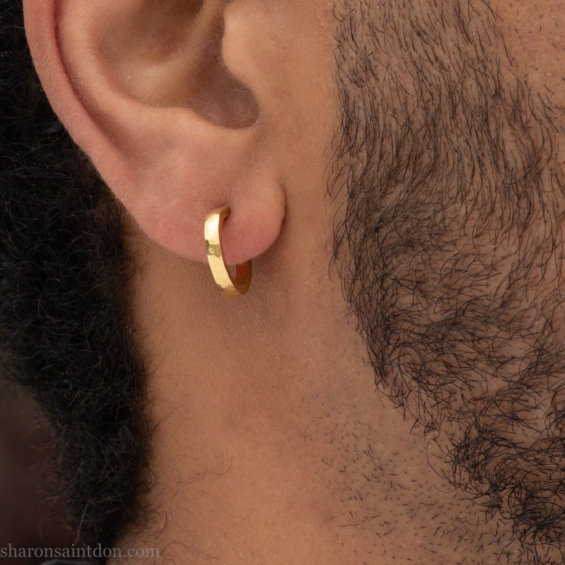 Hammered solid 18k yellow gold hoop earrings. 18mm diameter, 2mm wide, with a matte, brushed finish. Solid 18k gold posts and locking backs. Handmade by Sharon SaintDon in North America. Worn in photo by a young man with dark skin and black hair.