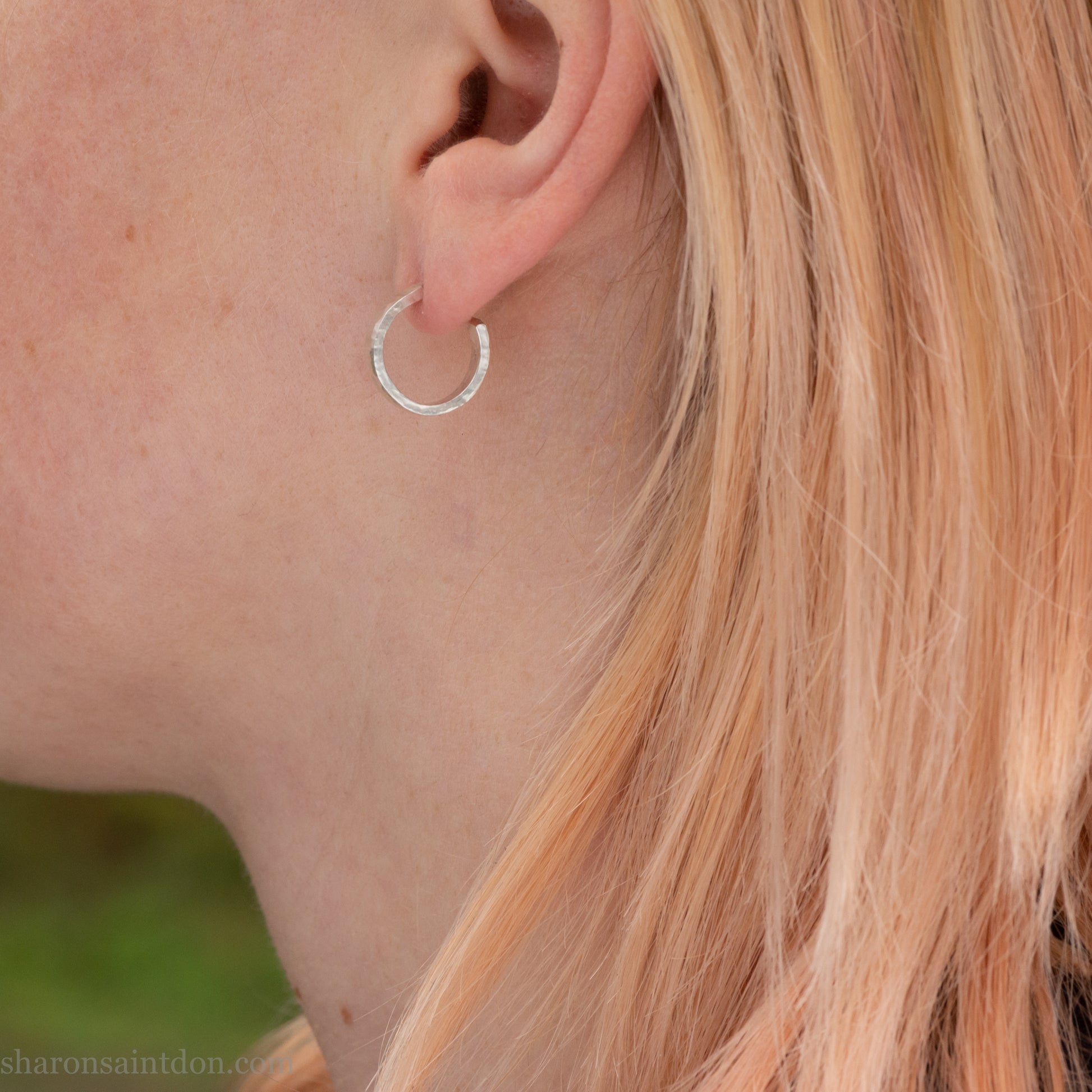 A close-up view of a person wearing handcrafted solid silver hoop earrings, 14mm diameter x 3mm wide x 1.5mm thick with a hammered matte finish.