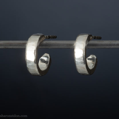 A pair of silver hoop earrings with a textured, hand-forged design, hanging on a metal bar against a dark background.