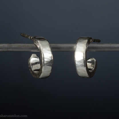 A pair of silver hoop earrings with a textured, hand-forged design, hanging on a metal bar against a dark background.