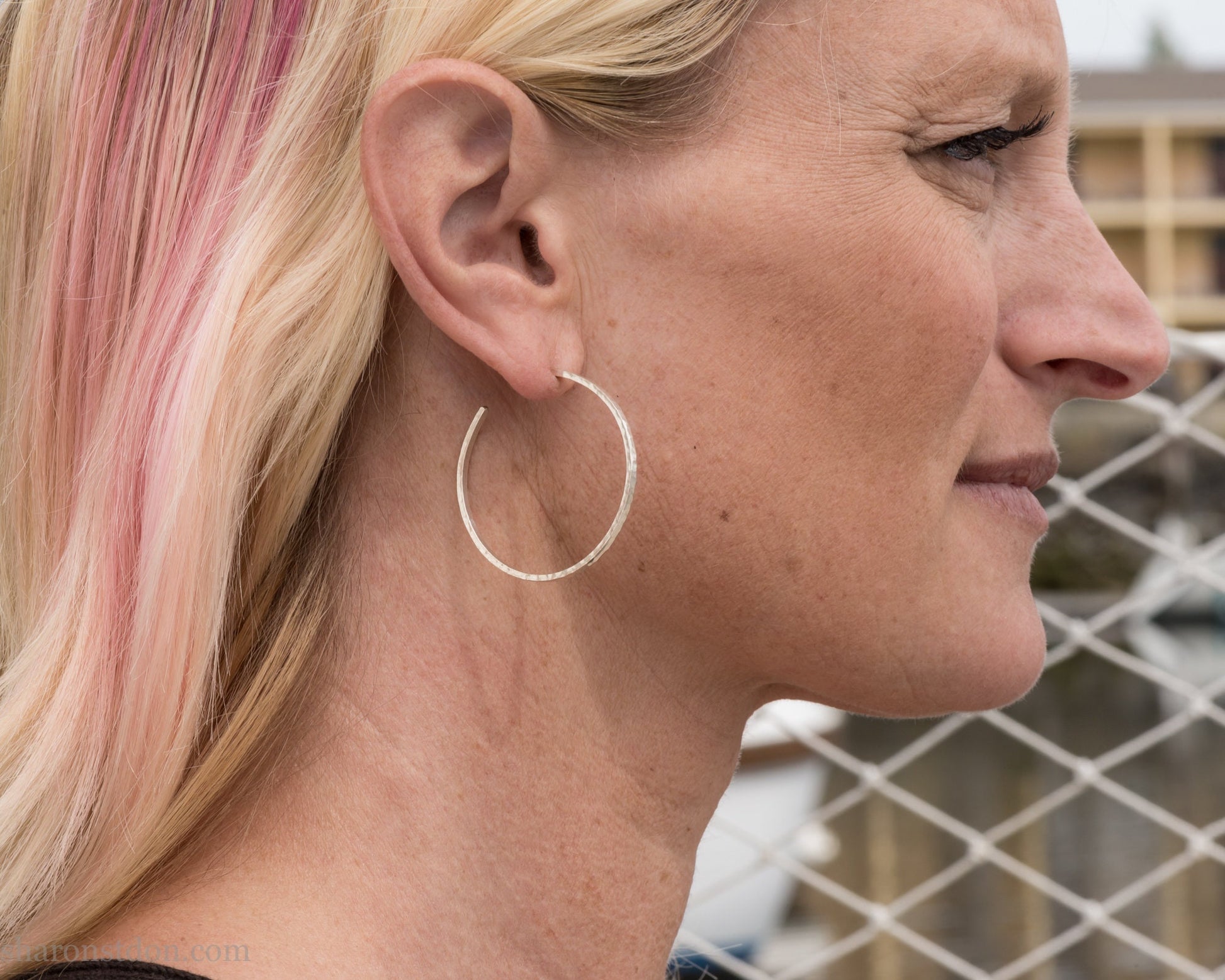 40mm diameter, 2mm wide, handmade 925 sterling silver hoop earrings. Comfortable, lightweight, daily wear hoop earrings with a hammered texture. Made by Sharon SaintDon in North America. Worn by a woman with light skin and blonde hair.