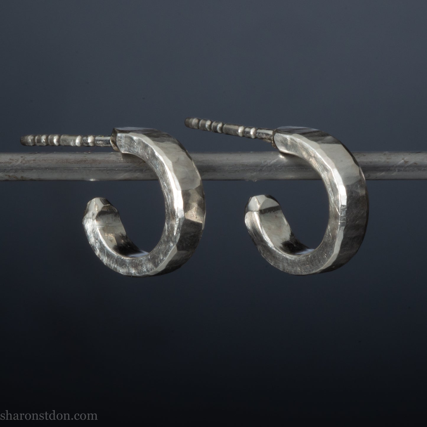 A pair of silver hoop earrings with a textured, hand-forged design, hanging on a metal bar against a dark background.