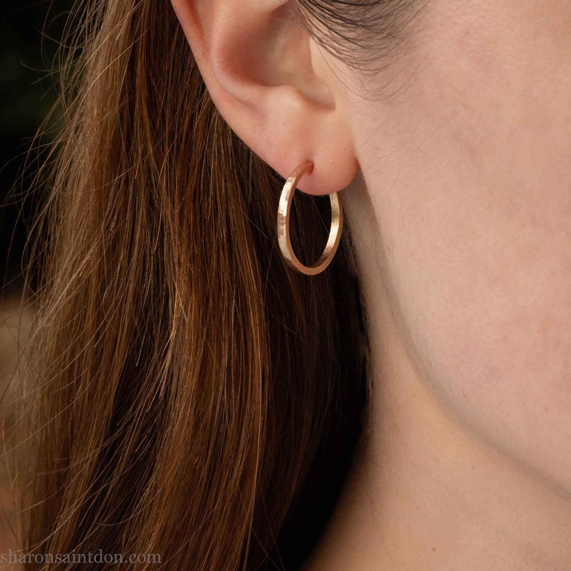 20mm diameter by 1.5mm thick  solid 14k yellow gold hoop earrings. Solid 14k gold posts and backs. Handmade by Sharon SaintDon in North America. Hammered from solid gold with a matte finish. Worn by a woman with brown hair and light skin.