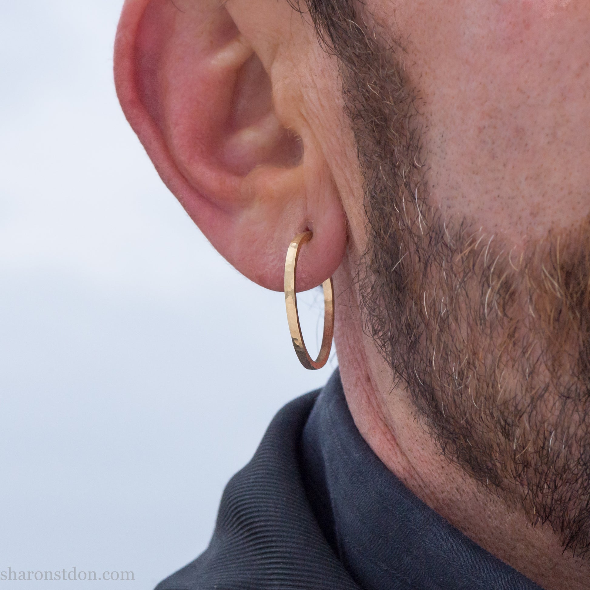 25mm diameter by 1.5mm thick handmade solid 14k yellow gold hoop earrings with solid 14k gold posts and backs. Handmade by Sharon SaintDon in North America. Hammered from solid gold with a matte finish. Worn by a man with light skin, dark beard and hair.