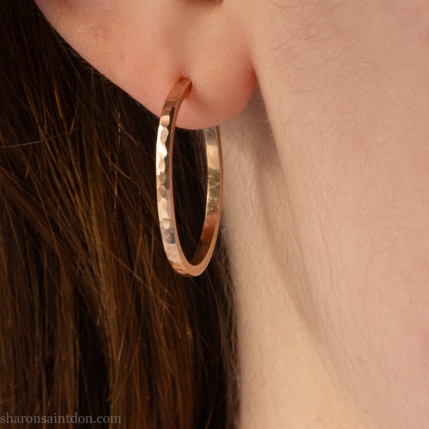 25mm diameter by 1.5mm thick handmade solid 14k yellow gold hoop earrings with solid 14k gold posts and backs. Handmade by Sharon SaintDon in North America. Hammered from solid gold with a matte finish. Worn by a woman with light skin and brown hair.
