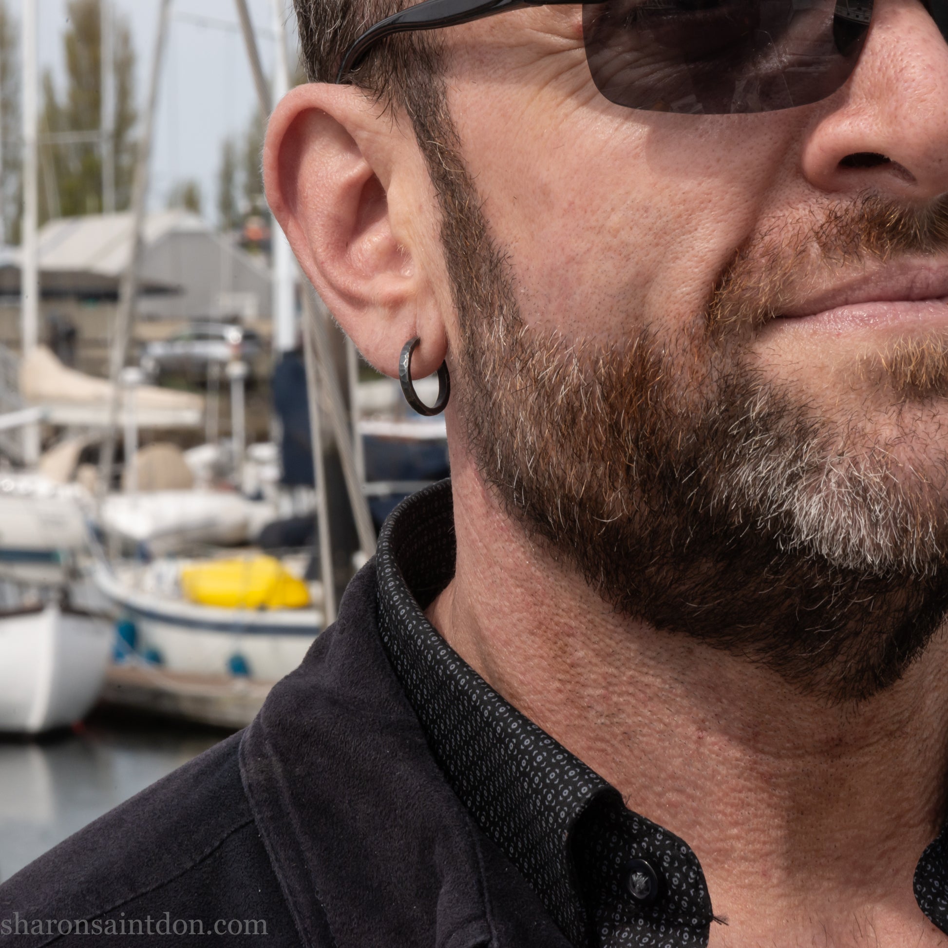 A close-up of a man's ear wearing a 18x2mm matte dark patina 925 sterling silver hammered hoop earring, with a blurred outdoor marina background.
