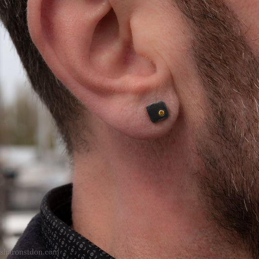 Handcrafted sterling silver square stud earrings with a 2mm canary yellow diamond set in the corner, hammered texture, oxidized black finish, shown worn on man's ear.