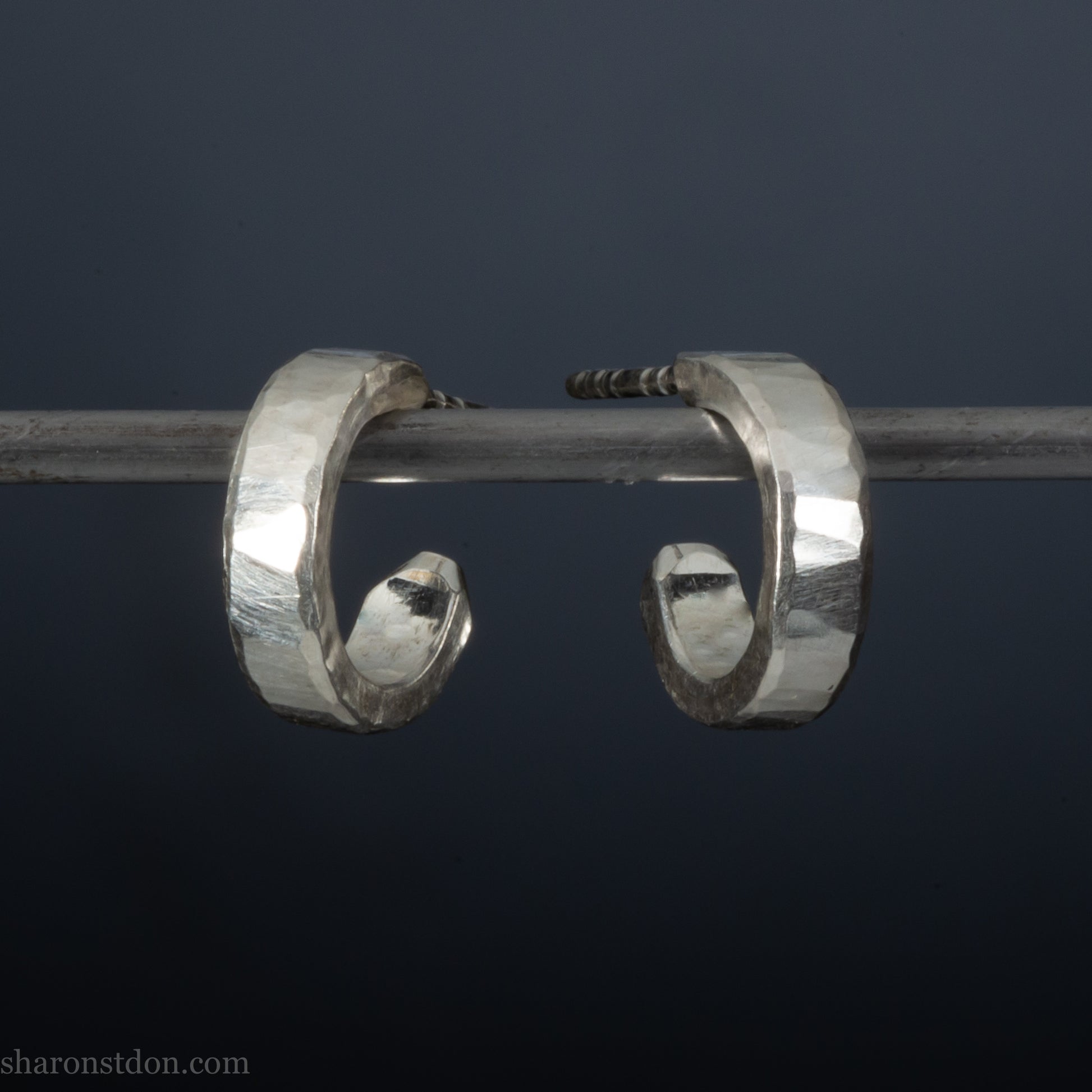 A pair of silver hoop earrings with a textured, weathered appearance, hanging from a metal hook against a dark background.