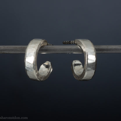 A pair of silver hoop earrings with a textured, weathered appearance, hanging from a metal hook against a dark background.