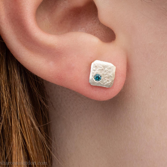 Square sterling silver and teal blue diamond stud earring, matte hammered finish, worn on ear.