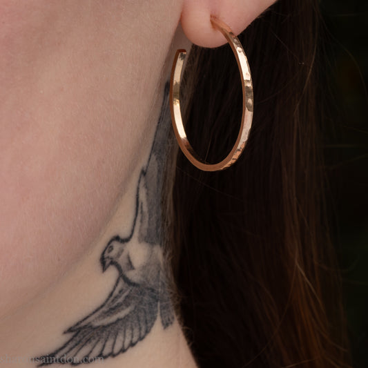 30mm diameter by 1.5mm thick, solid 14k yellow gold hoop earrings with locking posts and backs. Handcrafted by Sharon Saint Don in North America. Hammered from solid gold with a matte finish. Pictured on a woman with fair skin and dark red brown hair.