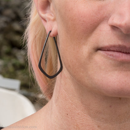 Person wearing hand hammered 925 sterling silver hoop earrings that are long, angular, teardrop shaped and oxidized black. SIZE: 5cm high x 32mm wide.