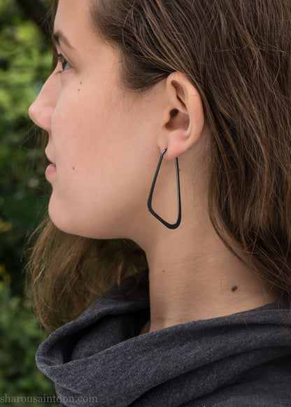 Person wearing hand hammered solid 925 sterling silver hoop earrings. Long, angular, low D shape, oxidized black, 5cm high x 32mm wide.