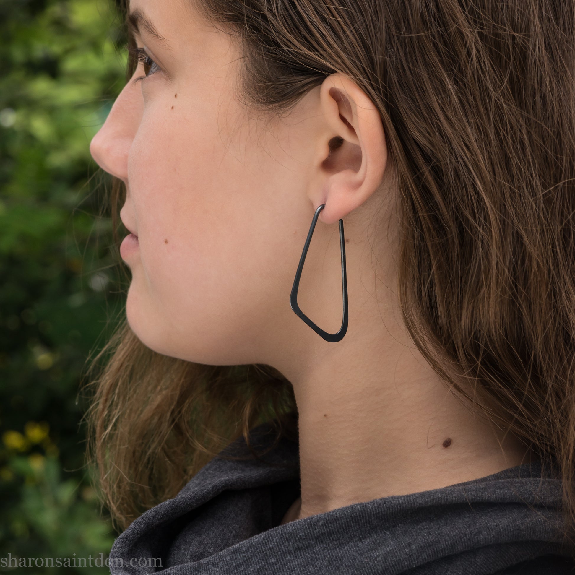 Person wearing hand hammered solid 925 sterling silver hoop earrings. Long, angular, low D shape, oxidized black, 5cm high x 32mm wide.