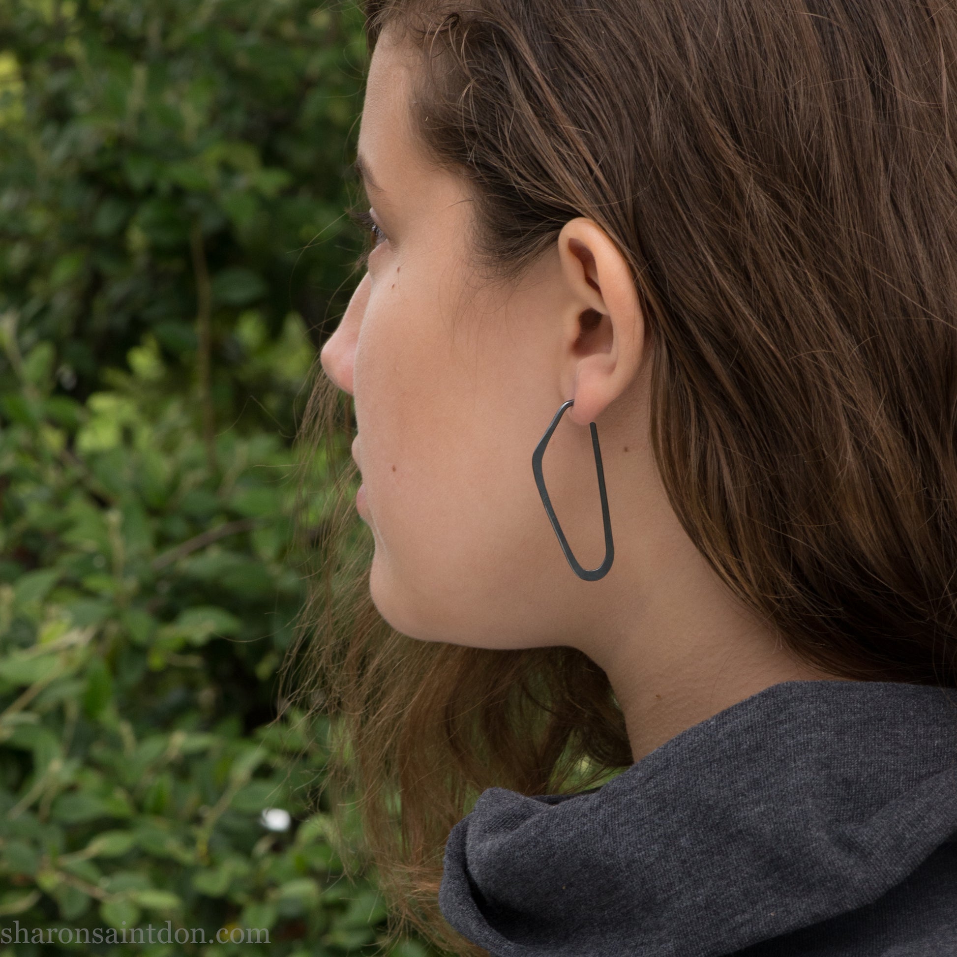 Person wearing hand hammered solid 925 sterling silver hoop earrings. Long, angular, high D shape, oxidized black, 47mm high x 19mm wide.