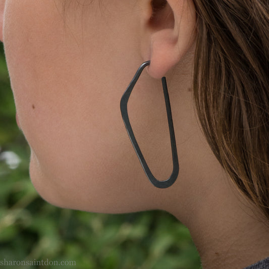 Person wearing hand hammered solid 925 sterling silver hoop earrings. Long, angular, high D shape, oxidized black, 47mm high x 19mm wide.
