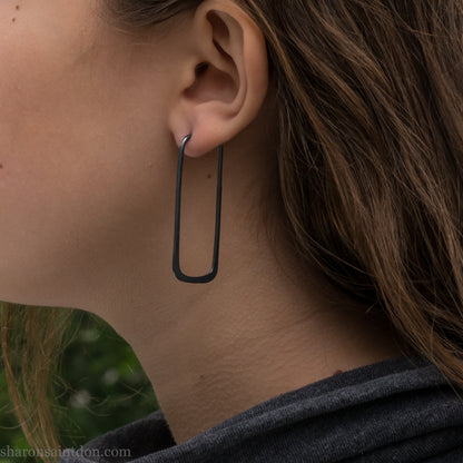 Person wearing hand hammered oxidized black rectangle hoop earrings in solid 925 sterling silver. 2x5cm, matte black patina.