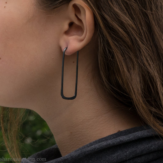 Person wearing hand hammered oxidized black rectangle hoop earrings in solid 925 sterling silver. 2x5cm, matte black patina.