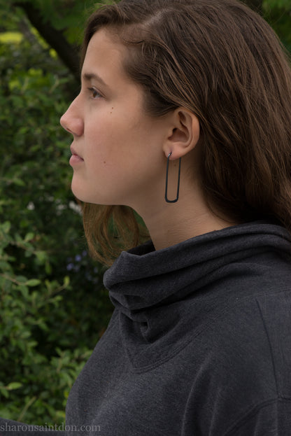 Person wearing hand hammered oxidized black rectangle hoop earrings in solid 925 sterling silver. 2x5cm, matte black patina.