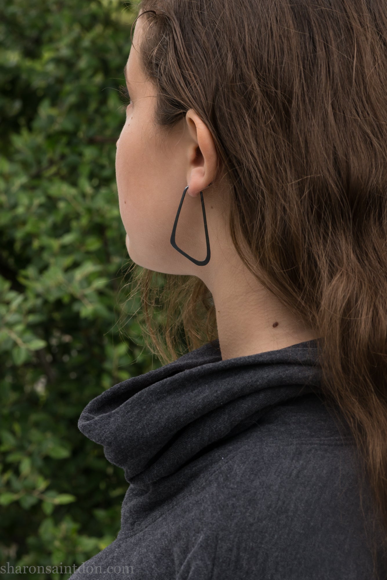 Person wearing hand hammered solid 925 sterling silver hoop earrings. Long, angular, low D shape, oxidized black, 5cm high x 32mm wide.