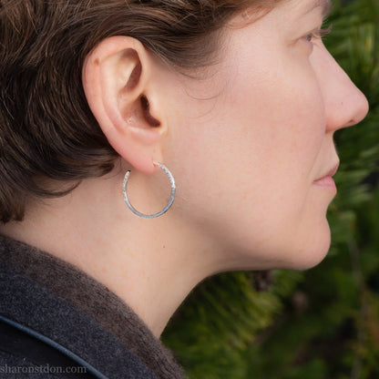 Hammered solid silver hoop earrings, handmade in the USA by Sharon Saint Don. Earrings are worn by a person with fair skin and short brown hair. 25mm diameter, 2mm wide, 1.5mm thick. solid silver posts and backs. Lightweight and comfortable for daily wear with a matte, brushed, shiny finish.