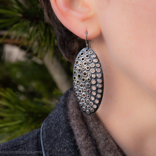 925 sterling silver dangle earrings, 50mm tall ovals with mesh holes punched by hand, and handmade ear wire french hooks. Oxidized black, dark patina rubbed back to shiny white shiny raised circles around the holes like little craters.