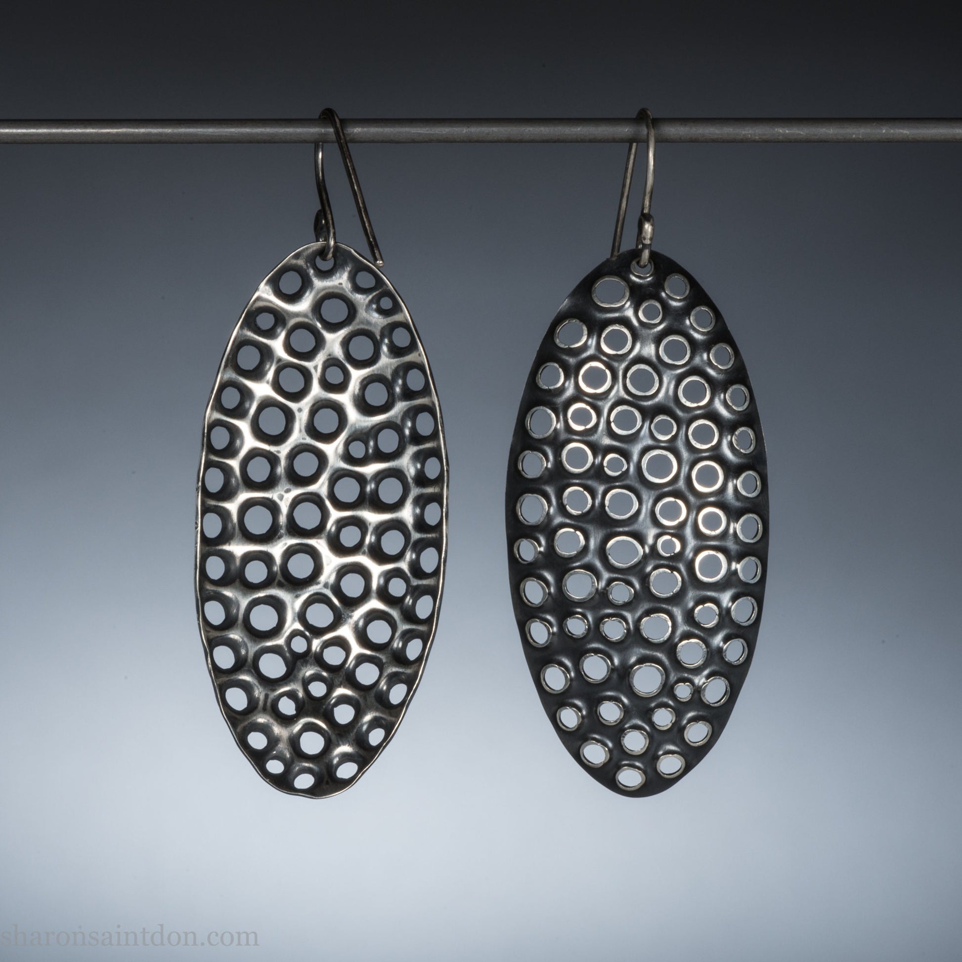 925 sterling silver dangle earrings, 50mm tall ovals with mesh holes punched by hand, and handmade ear wire french hooks. Oxidized black, dark patina rubbed back to shiny white shiny raised circles around the holes like little craters.